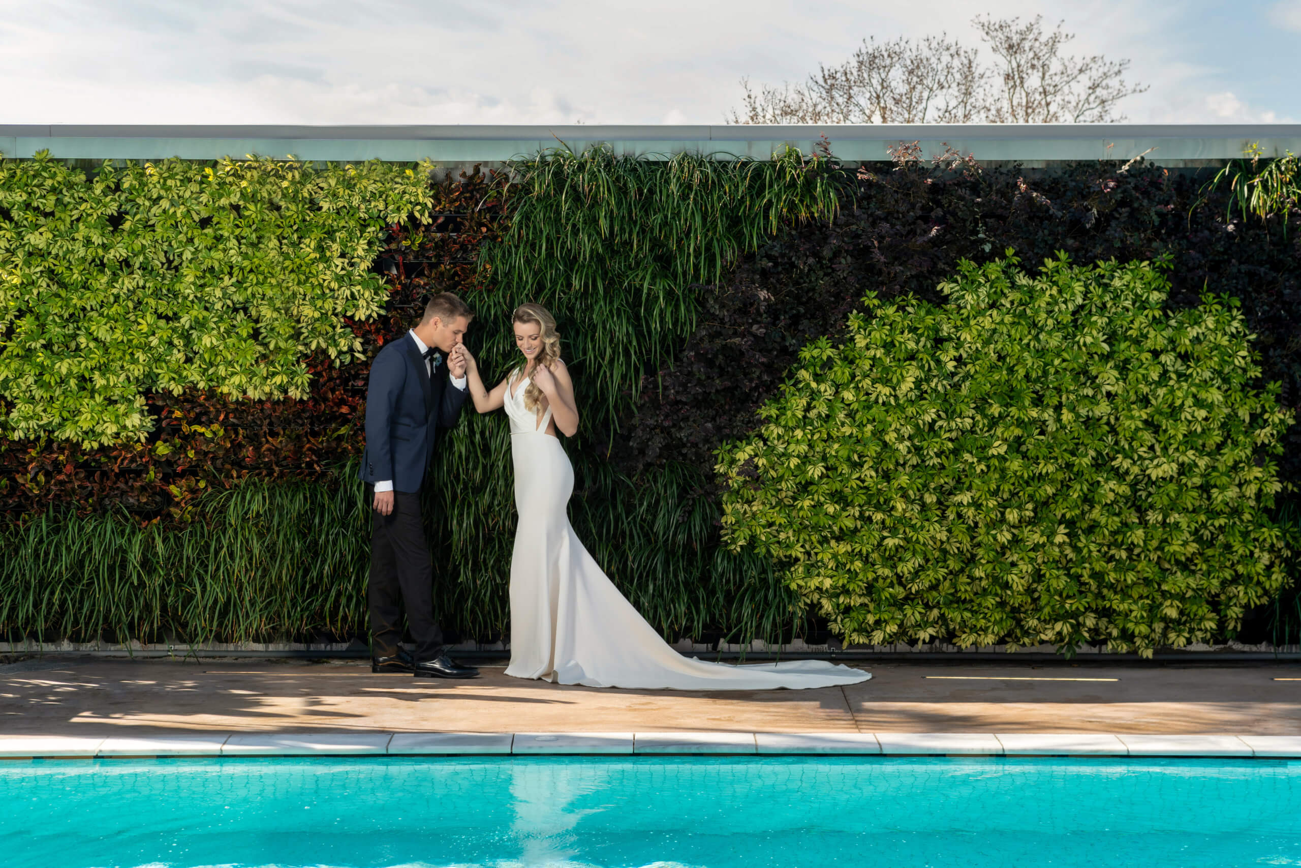 Una novia con un vestido largo blanco y un novio con un traje oscuro posan junto a una piscina azul, y el novio le besa la mano a la novia. Están frente a un exuberante jardín vertical de color verde.