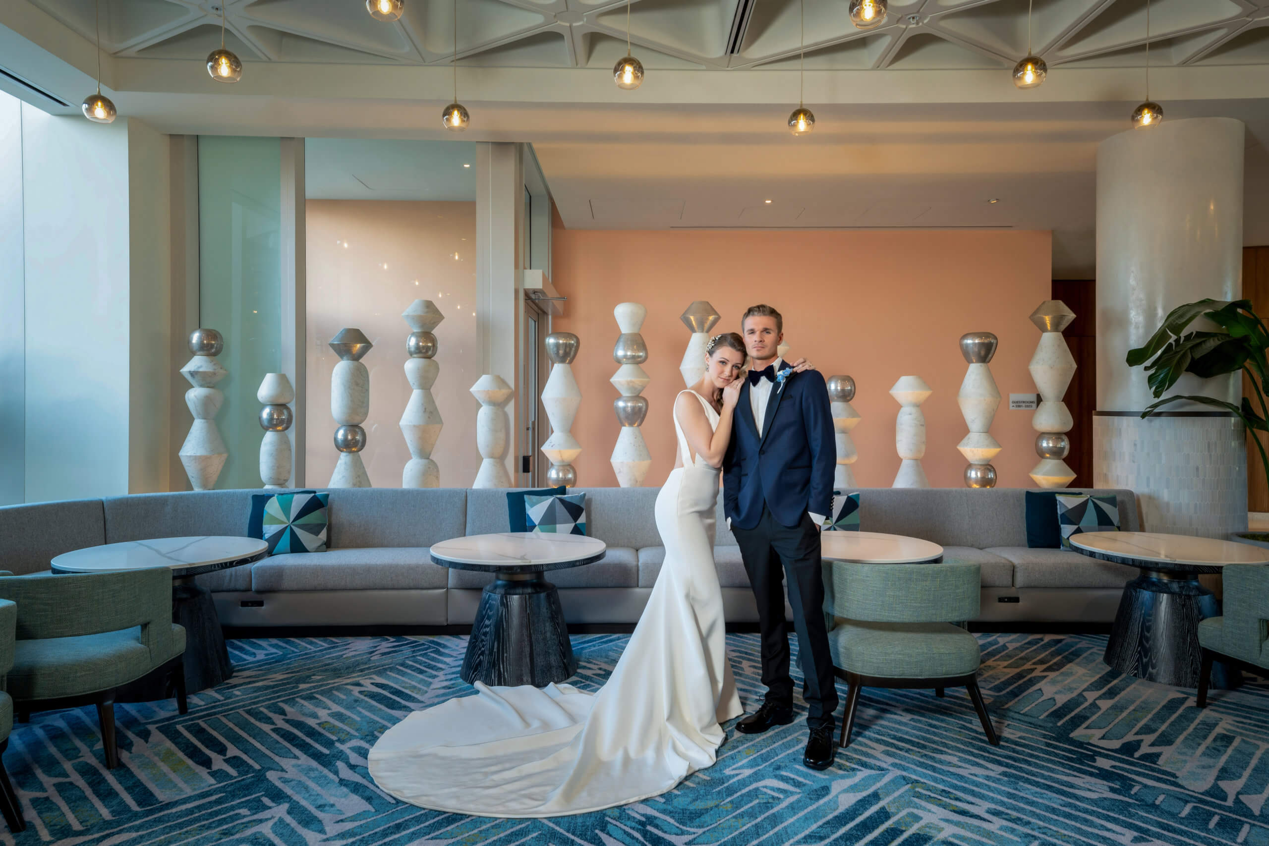 Una novia con un vestido blanco y un novio con un traje oscuro posan juntos en un moderno « lounge » con decoración geométrica, sofás grises, mesas redondas y una alfombra azul estampada. En el fondo se alinean esculturas esféricas y cilíndricas.