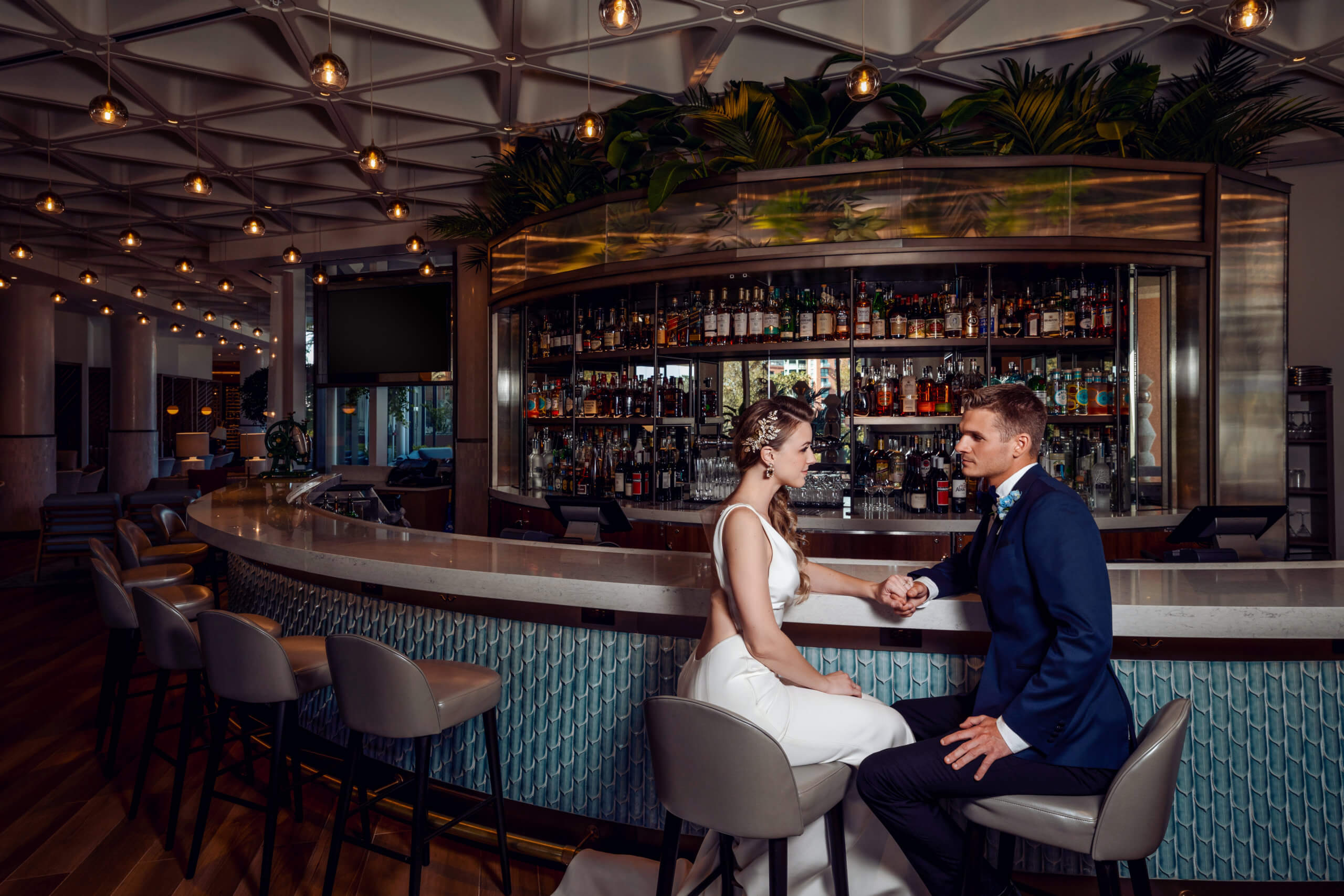 A bride and groom sit at a modern bar, holding hands and facing each other, dressed in formal wedding attire. The bar is stylish with plants, pendant lights, and a wide selection of bottles behind them.