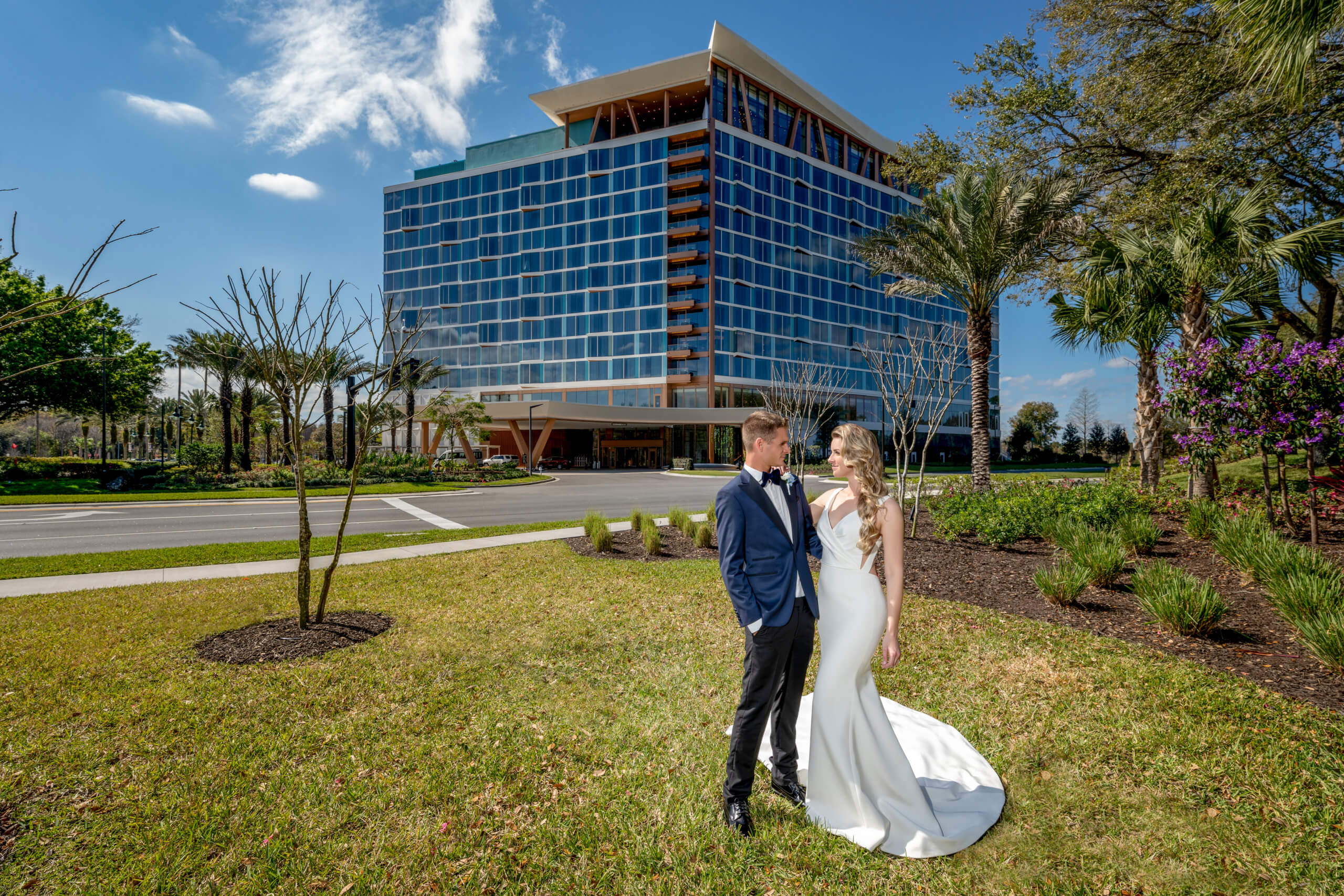 A bride and groom stand together on a lawn, gazing at each other, with a modern glass hotel building and palm trees in the background under a bright blue sky.