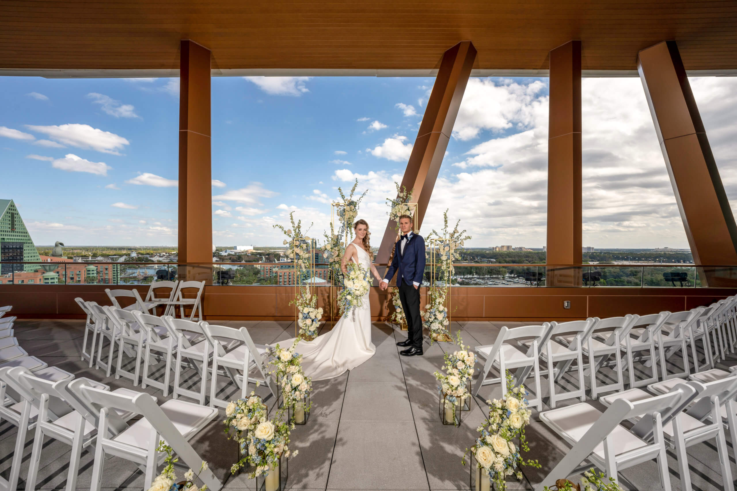 A bride and groom stand together at an outdoor rooftop wedding ceremony, surrounded by white chairs and floral arrangements, with a cityscape and blue sky visible in the background.