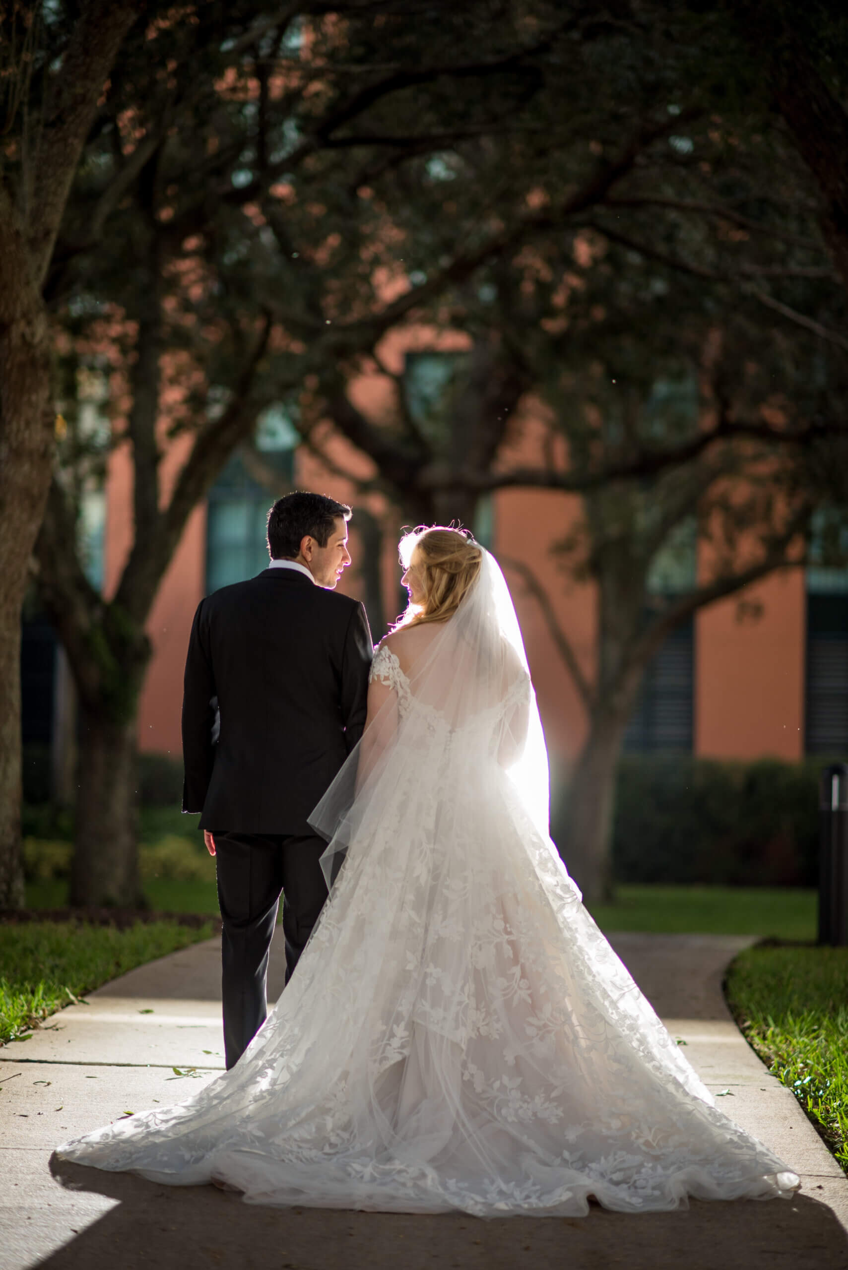A bride in a white wedding gown and veil stands beside a groom in a black suit, holding hands and looking at each other on a sunlit path surrounded by trees.