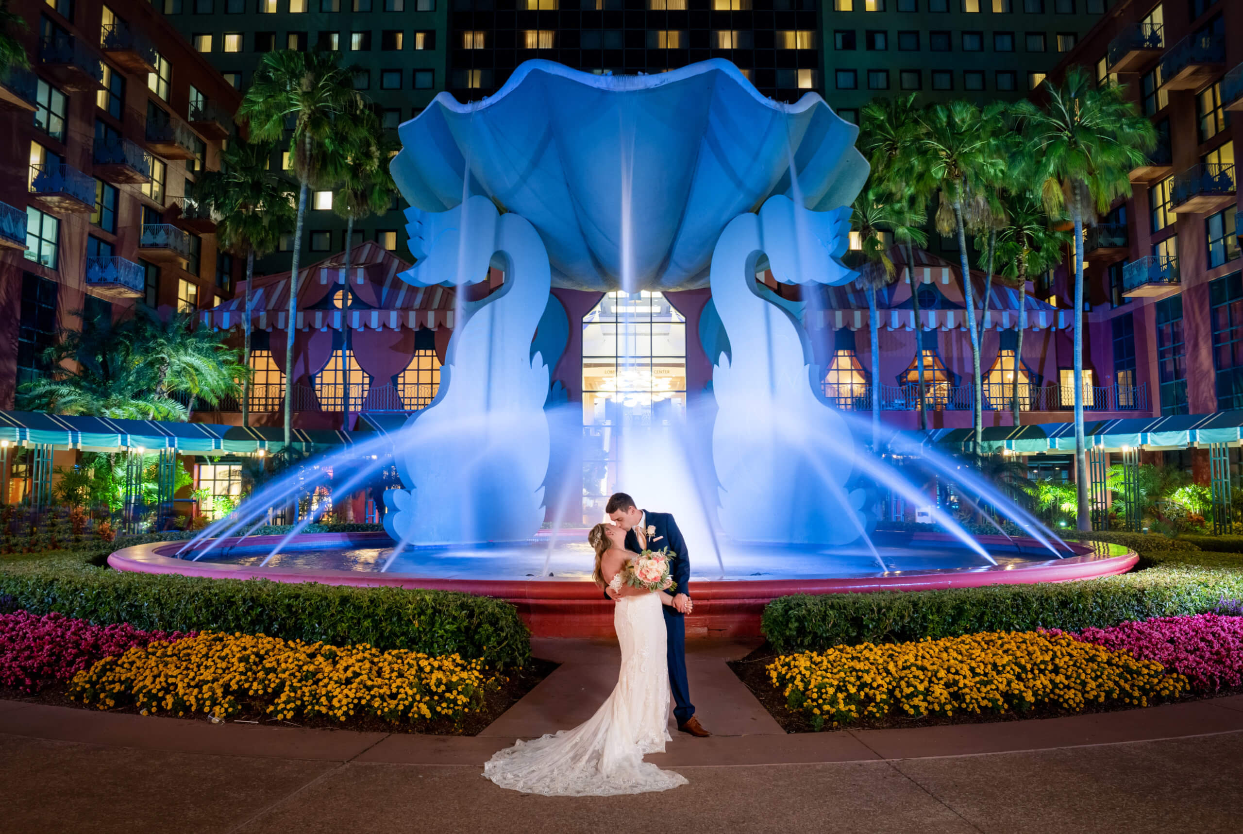 A bride and groom kiss in front of a large, illuminated fountain shaped like a seashell with swan sculptures, surrounded by colorful flowers and palm trees, at night.