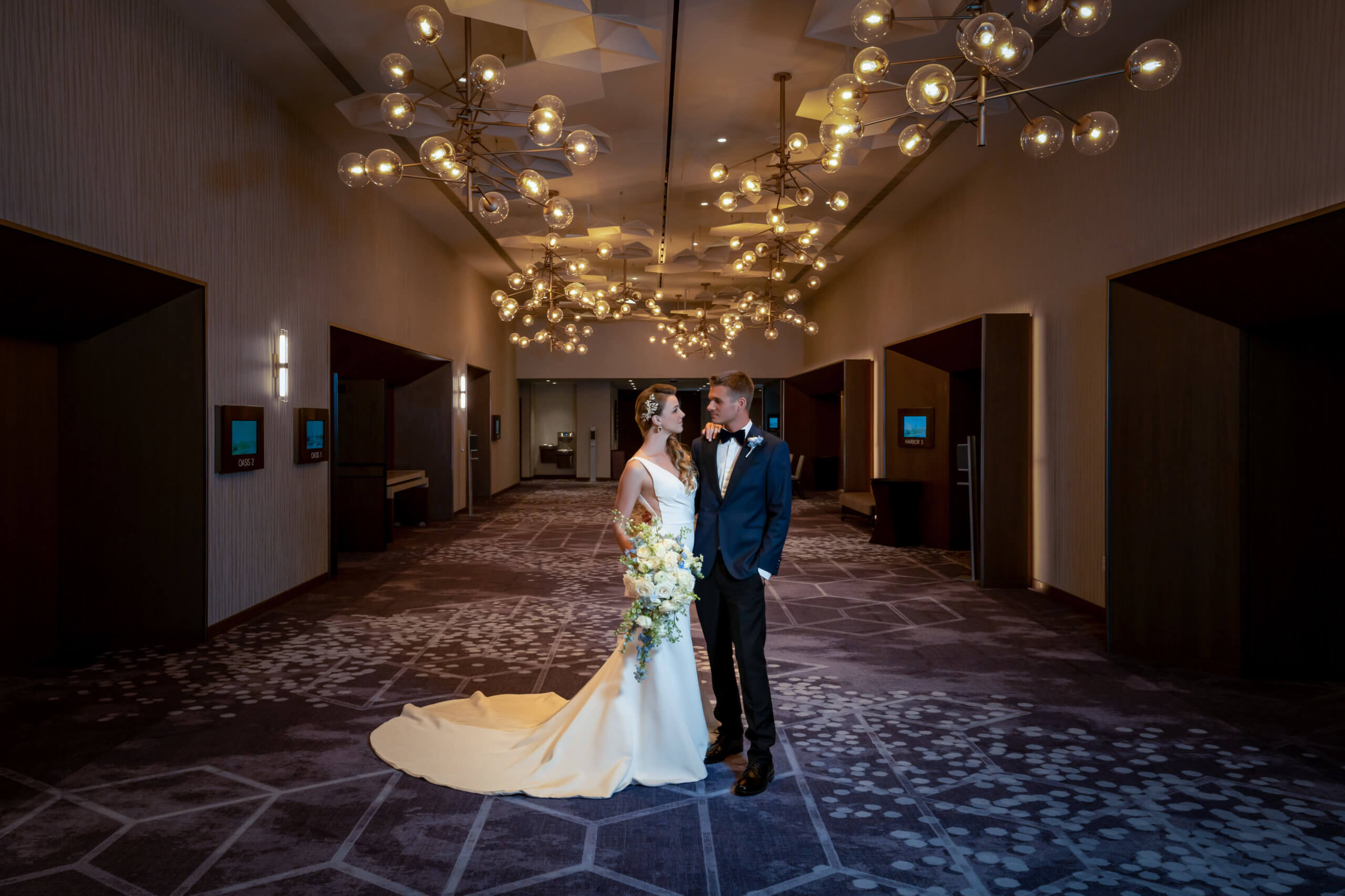 A bride in a white gown and a groom in a dark suit stand together in an elegant, modern hallway with geometric carpet and glowing, contemporary chandeliers overhead.