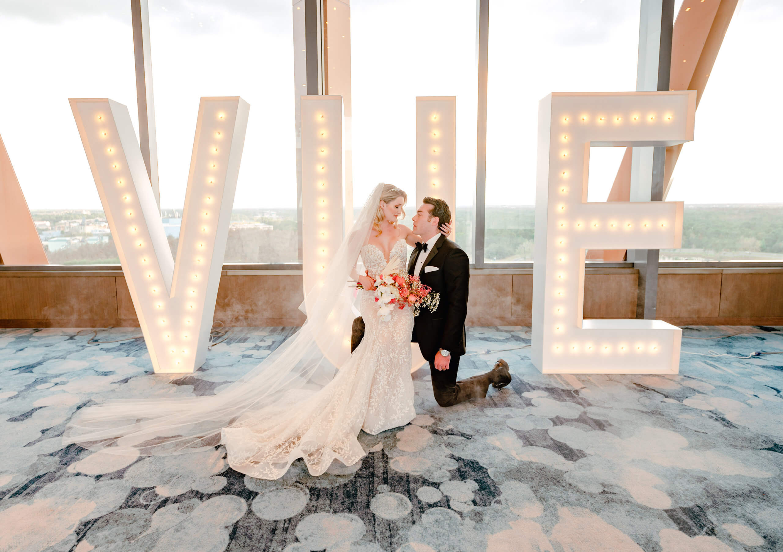 「VUE」と書かれた大きなイルミネーションの文字の前で、新郎新婦が並んでひざまずき、互いを見つめ合っている。花嫁はブーケを手にし、長いベールをまとっており、二人の背後の高い窓からは街並みが望める。