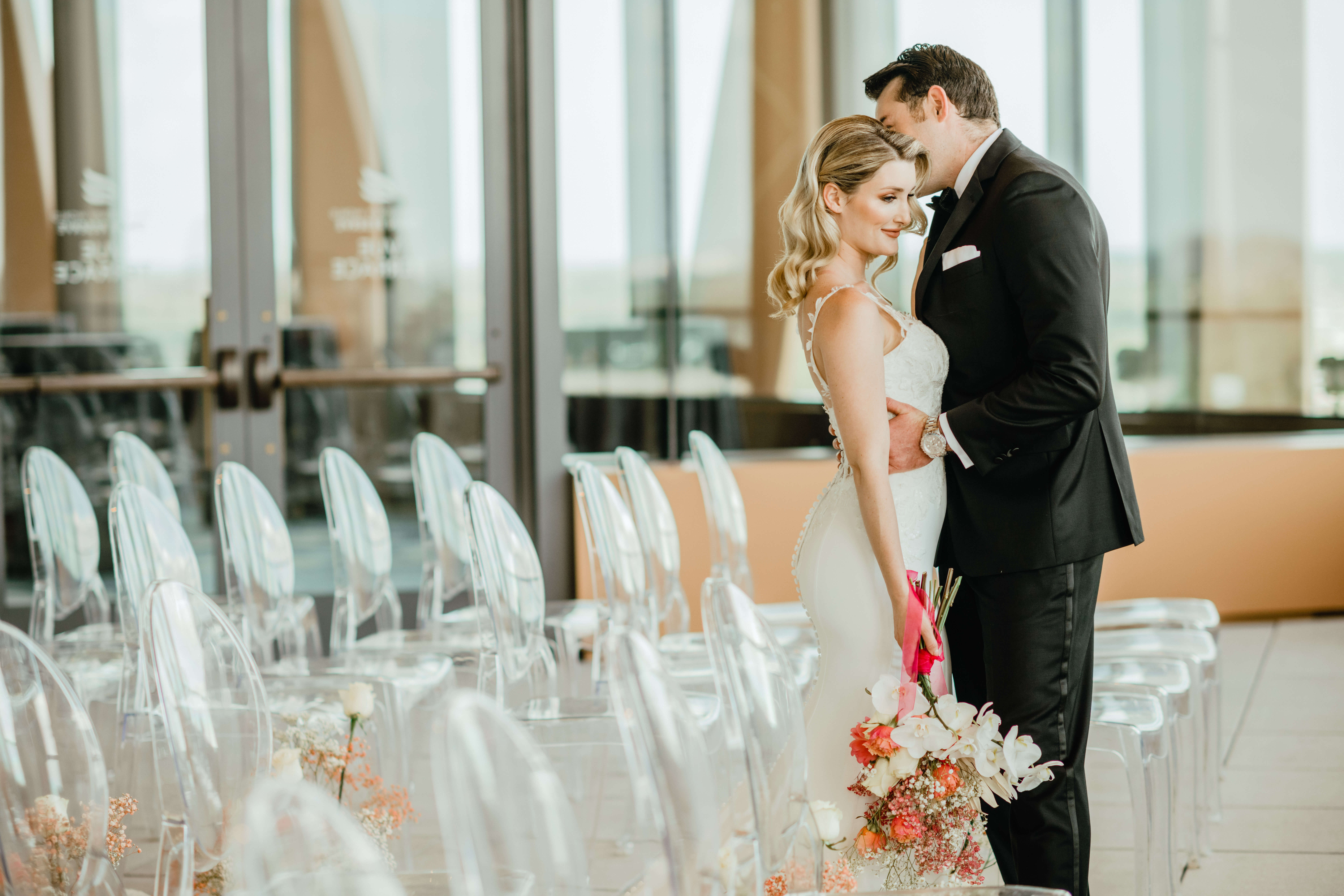 A bride and groom embrace tenderly in an elegant indoor venue with transparent chairs and floral arrangements. The bride holds a bouquet, and both look content and joyful.