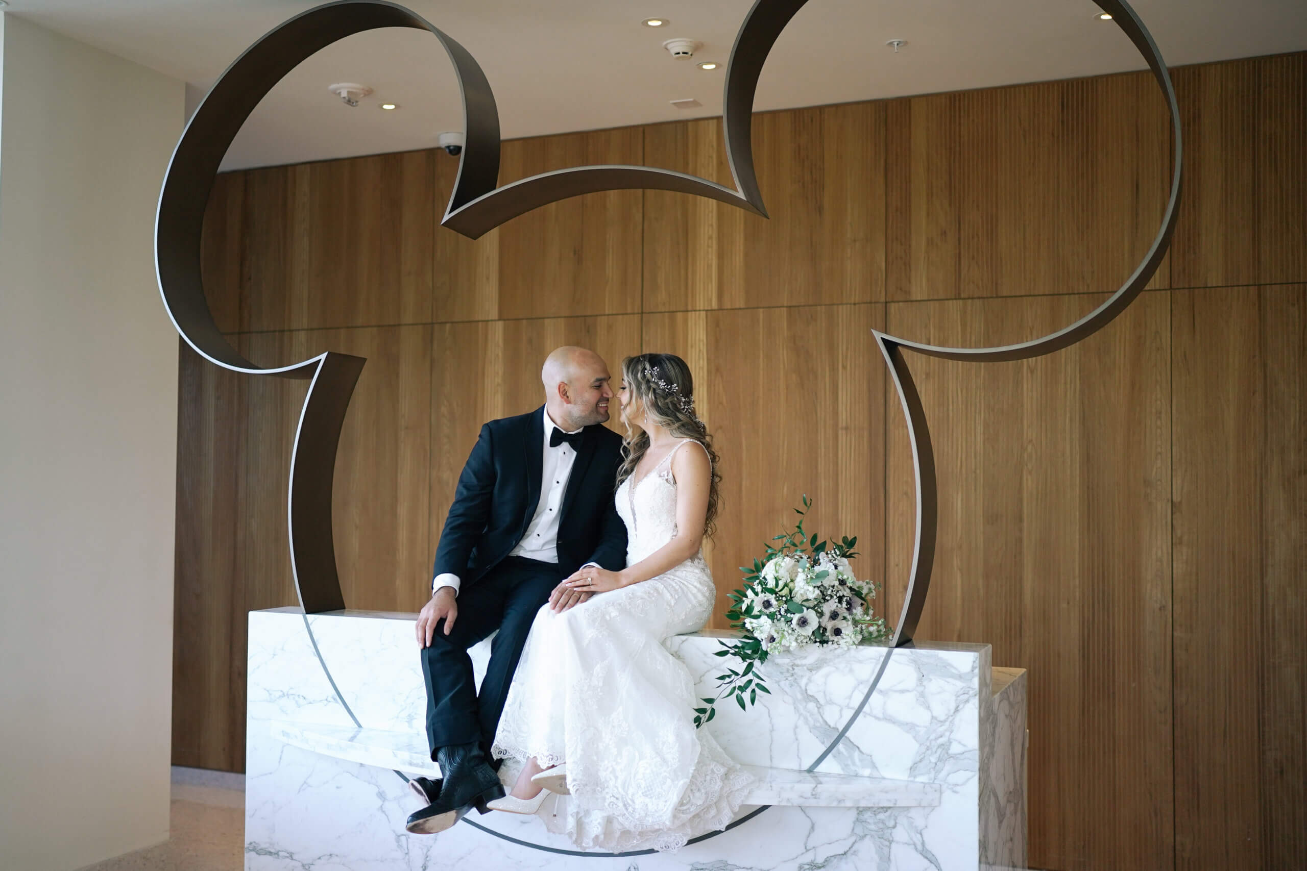A bride and groom sit together on a marble platform, surrounded by a large metal outline of Mickey Mouses head, gazing at each other lovingly in an elegant, wood-paneled room.