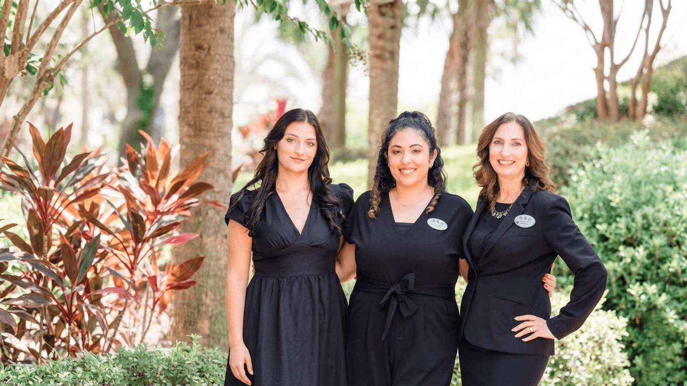 Trois femmes vêtues de robes noires et portant des badges nominatifs se tiennent à l'extérieur ; elles sourient et posent ensemble devant des arbres et une végétation luxuriante par une journée ensoleillée.